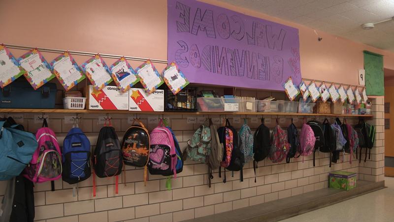 Image of backpacks hanging from hooks in the hallway of an elementary school.